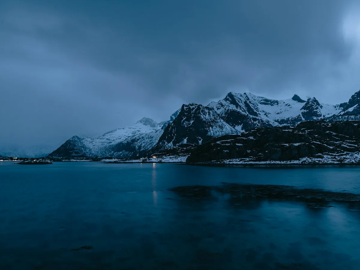 A dusk view of snow-covered Arctic mountains above a still fjord, in deep blue winter light
